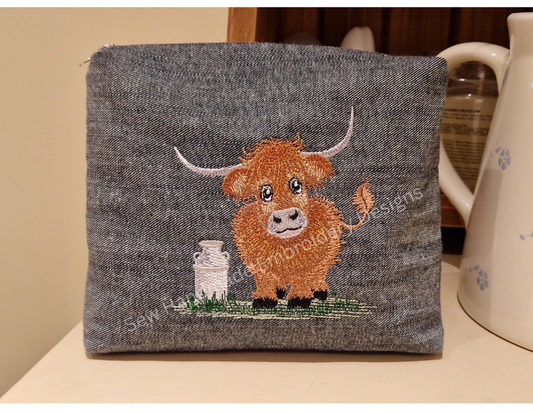 Machine embroidery design of a cute & fluffy Highland cow with a creamery can standing on a patch of green grass stitched onto a denim zipper bag / pouch - instand digital download